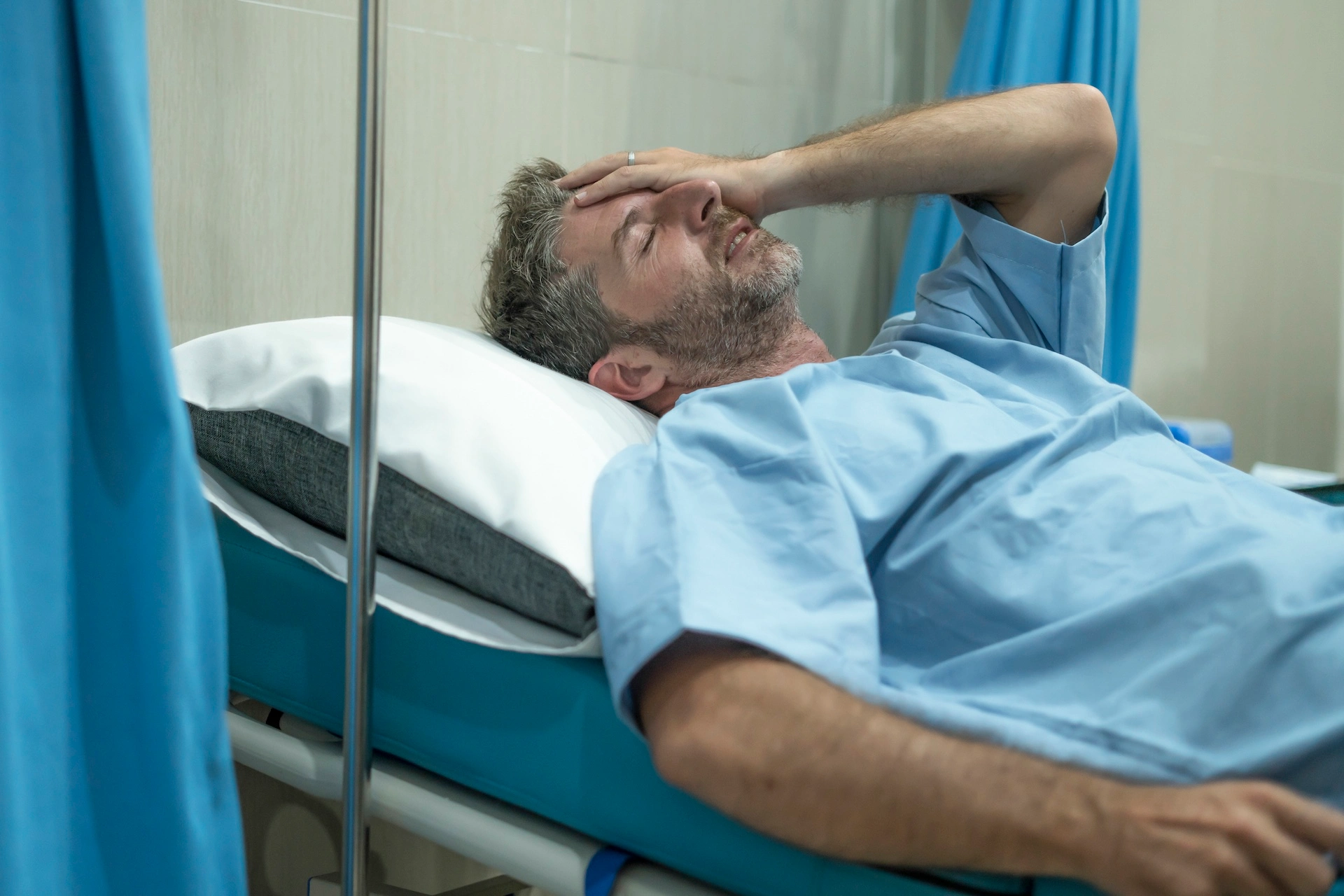 Older man lying on a hospital bed in an emergency room holding his head in pain.