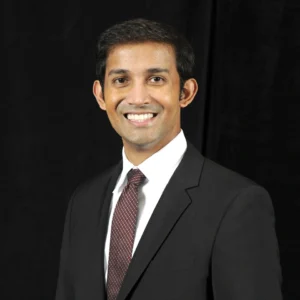 Dr. Saxena wearing a black suit jacket, white shirt, and maroon tie.