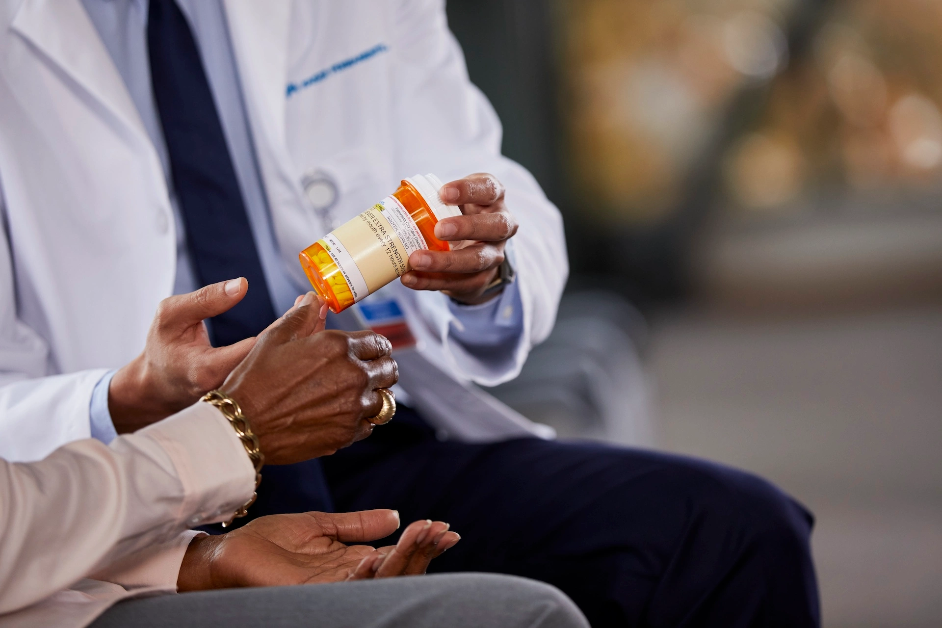 pharmacist seated in white coat holding a pill bottle and handing it to a seated patient.