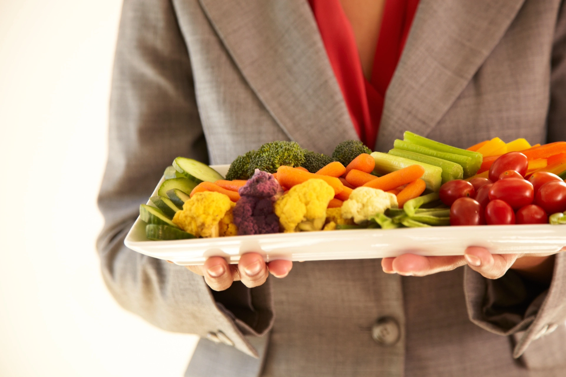 woman wearing a red shirt and brown suit jacket holding a plate of raw vegetables